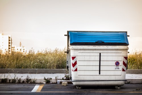 A large, white plastic commercial rubbish skip with a blue hinged lid, positioned on an asphalt surface beside a pavement. The skip features red and white chevron reflective markings on its sides and a small circular no parking sign attached to the front. Surrounding the skip, there is overgrown grass and weeds at the edge of the pavement, with some small stones and debris visible on the ground nearby. In the background, a distant view of a clear sky and faint outlines of buildings or industrial structures can be seen, suggesting an urban or suburban environment. The scene is illuminated by natural daylight, emphasizing the texture and colour of the skip, which appears sturdy and utilitarian, suitable for waste collection and rubbish removal services provided by companies such as Garden Clearance Palmers Green, operating within the Palmers Green area in North London.